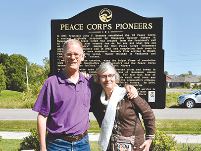 Daniel Grundtner in purple shirt with Ruth Alliband in white scarf in front of Peace Corp sign