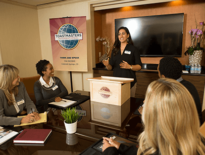 Woman standing behind lectern speaking to people around table
